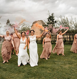 A joyful bridal party with colored smoke bombs creating a whimsical backdrop, all wearing matching dresses, with the brides in the center