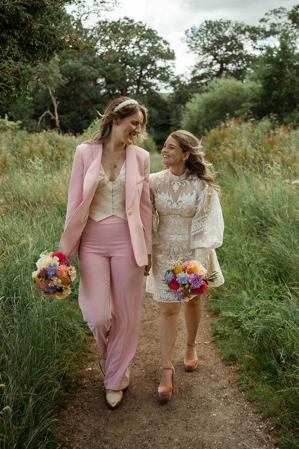 Lesbian couple on their wedding day walking hand in hand smiling at each other with colourful bouquets captured by Feminist & LGBTQ+ wedding photographer, Rebel Love Club