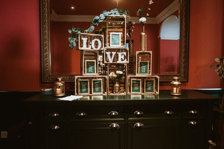Wedding venue lobby area, decorated with gold boxes, photo frames and LOVE letters. All standing on a black cabinet with a gold mirror behind and red walls. Style and decor by 4yaparty Wedding & Events, an LGBTQ+ supplier, part of the Rebel Love Directory. 