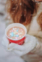 Gloved hands hold a red cup of hot chocolate with colorful marshmallows. Snowflakes rest on brown hair, creating a cozy, wintery feel.