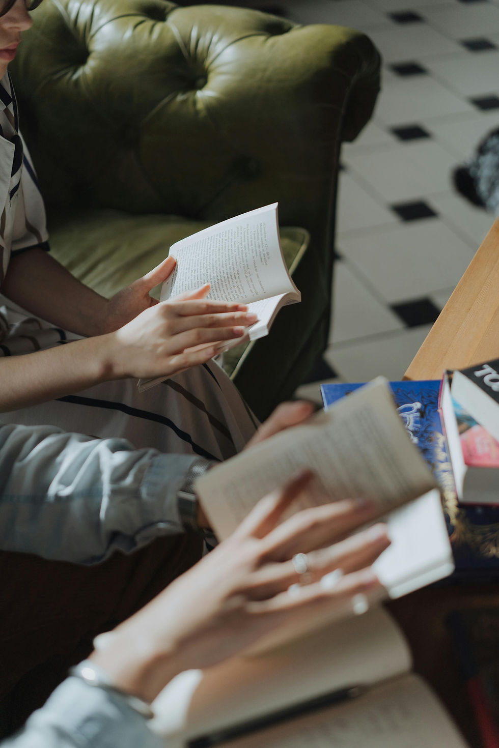 Hands holding open books, sitting on a green sofa. Books on a wooden table. Tiled floor background. Relaxed, focused atmosphere.
