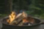 Log fire burning in a round metal pit outdoors, with smoke rising against a blurred forest background, creating a cozy atmosphere.