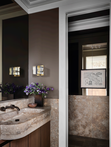 A powder room featuring Italian Travertine for the stone countertop and walls. The bathroom is accented with modern wall-mounted light fixtures, a sleek mirror, and fresh flowers on the countertop. The space exudes elegance with neutral tones and minimalistic design.
