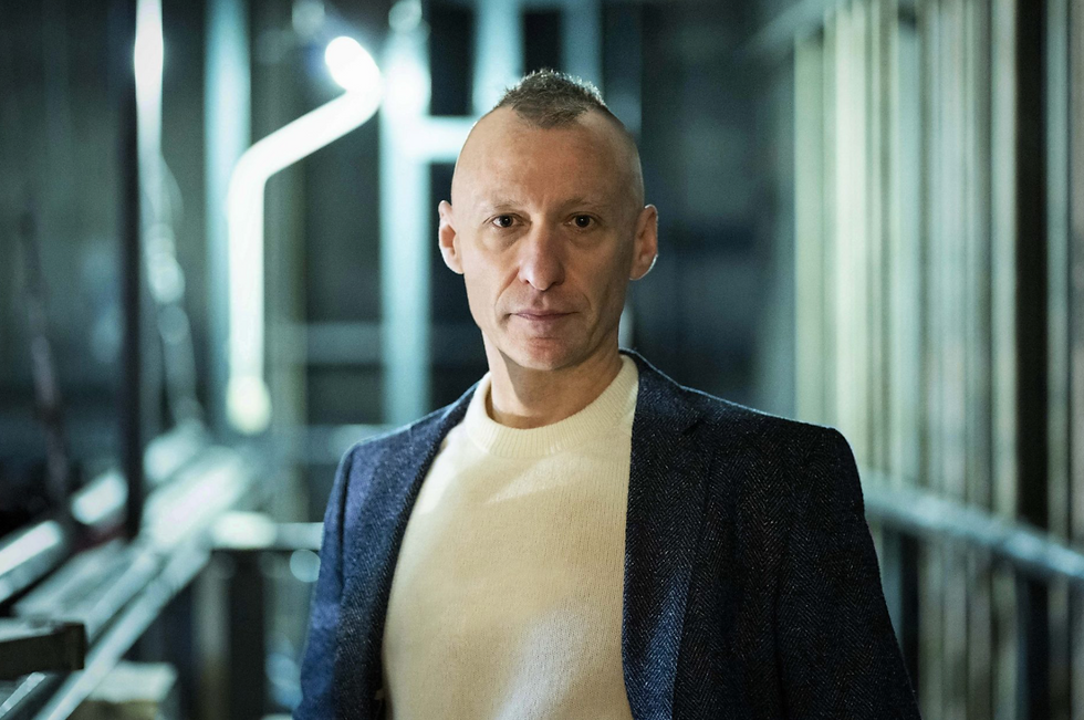 The image features Sergio Blanco, a renowned playwright, standing in an industrial or backstage environment. He has a serious, contemplative expression on his face, looking directly at the camera. Blanco has a distinct appearance with a closely shaved head and is wearing a dark blazer over a light-colored sweater. The background is blurred, emphasizing Blanco as the focal point, and includes structural elements like pipes and beams, possibly indicating a theater's backstage area or a similarly industrial setting. The overall mood of the image is introspective and professional, reflecting Blanco's influential presence in the world of contemporary theater.