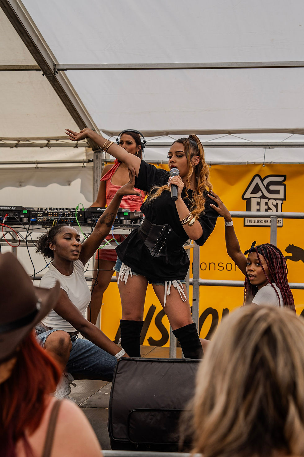 Female singer performs on stage with dancers under a tent, holding a microphone, with an AG Fest banner behind her.