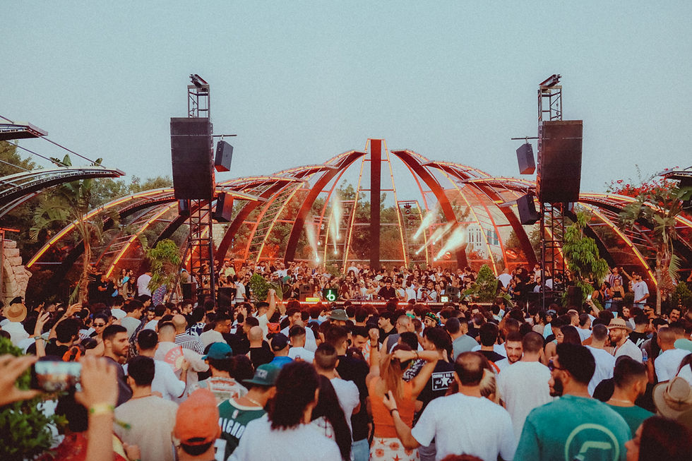 Crowd enjoying a vibrant outdoor concert under a red-arched structure, with colorful lights and lush greenery. Festive and energetic atmosphere.