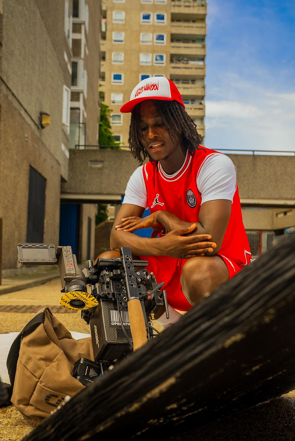 Young man in red sports attire adjusts a camera outdoors, wearing a red and white cap. Urban setting with buildings in the background.