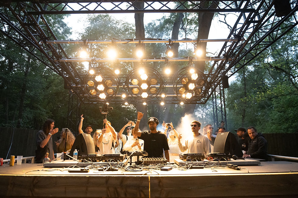 DJ performing outdoors under bright lights, surrounded by people celebrating. Trees and smoke create an energetic atmosphere.