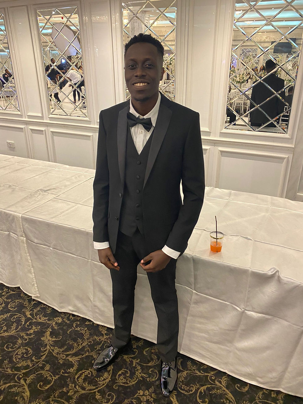 Micheal Dolo in a black tuxedo smiling, standing in an elegant room with mirrored walls and ornate carpet. A drink is on the table beside him.