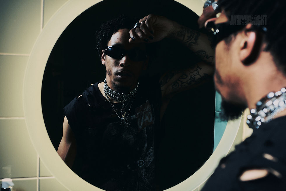 Man in sunglasses and layered necklaces poses in front of a circular mirror, displaying tattoos. Reflected in bathroom setting, edgy style.
