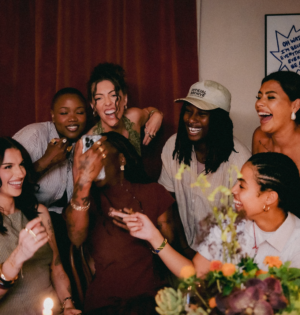 Group of friends laughing and posing for a selfie at a warm indoor party, with flowers on the table and a poster with text in back.