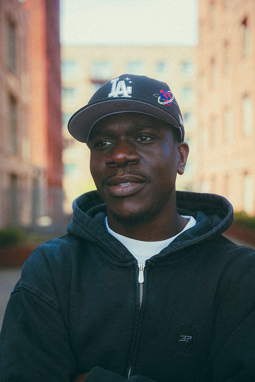 Man in an LA cap and black hoodie stands outdoors against blurred apartment buildings, looking thoughtful.