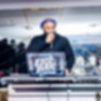 DJ in blue headphones speaks into a mic behind a laptop labeled ESCO BAR at a lively indoor party.