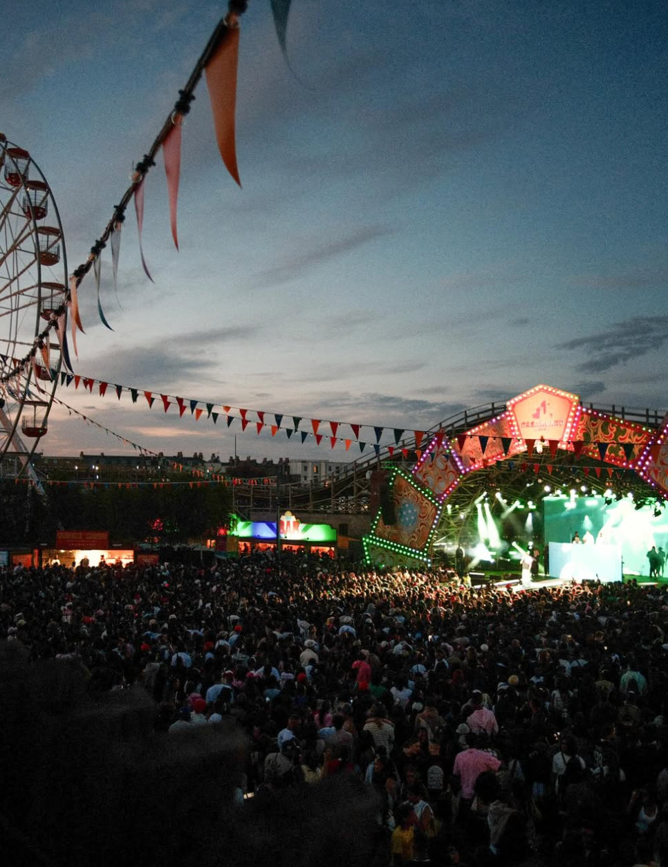 Crowded music festival at dusk with colorful bunting and a lit stage. Ferris wheel and buildings in the background, festive atmosphere.