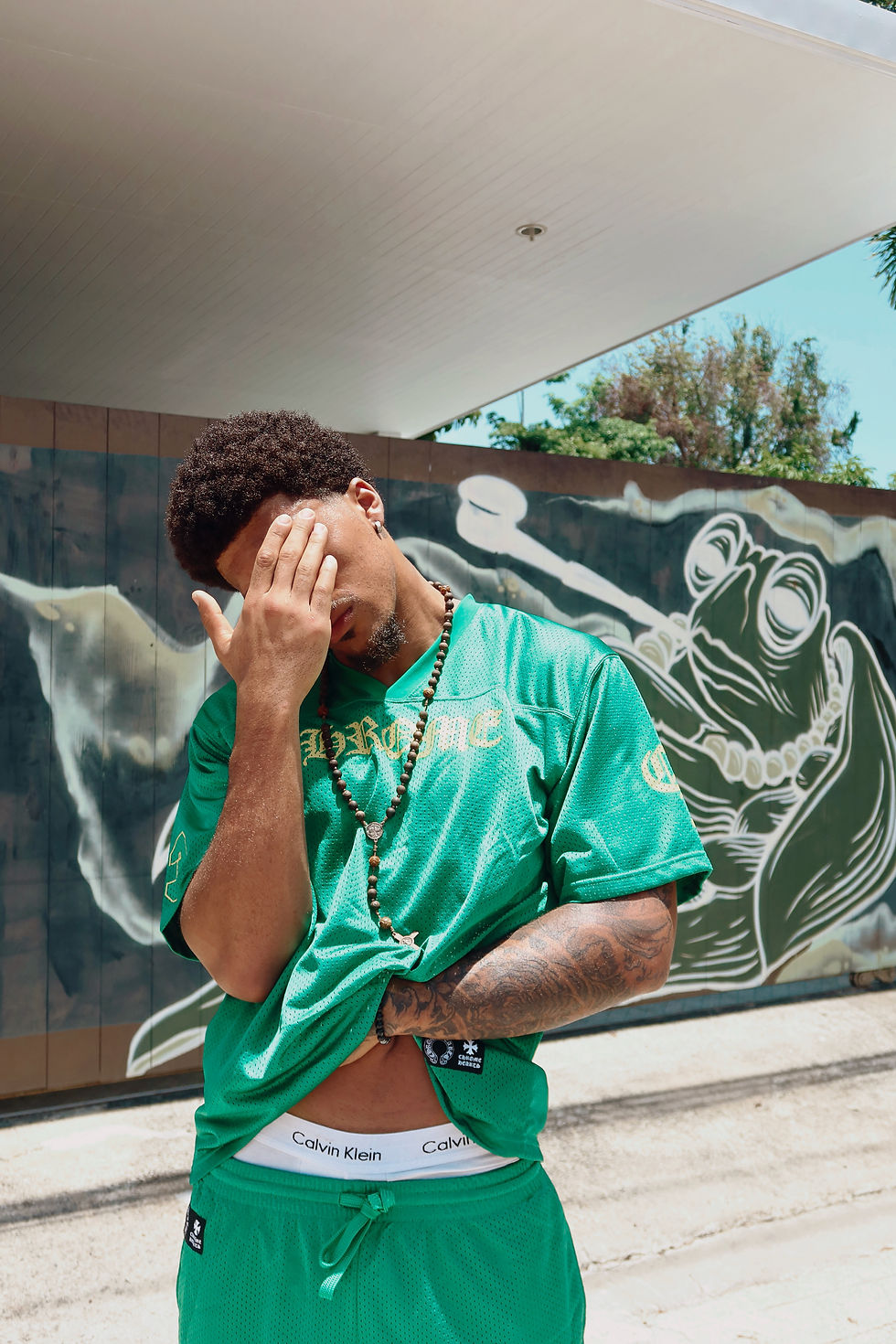 Man in green shirt and shorts covers face with hand in front of frog mural, showing Calvin Klein waistband. Sunny outdoor setting.