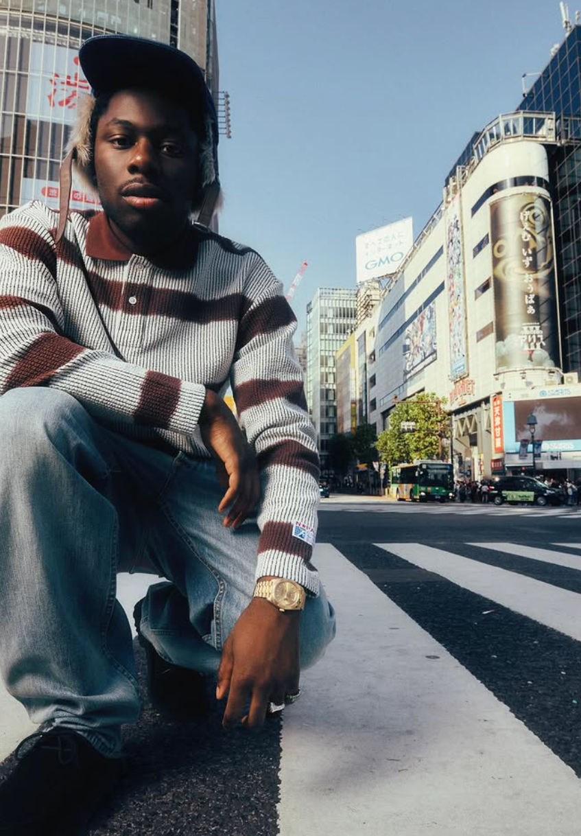 Young man kneeling on a city street, wearing a striped sweater and cap. Urban background, with vibrant billboards and clear sky.