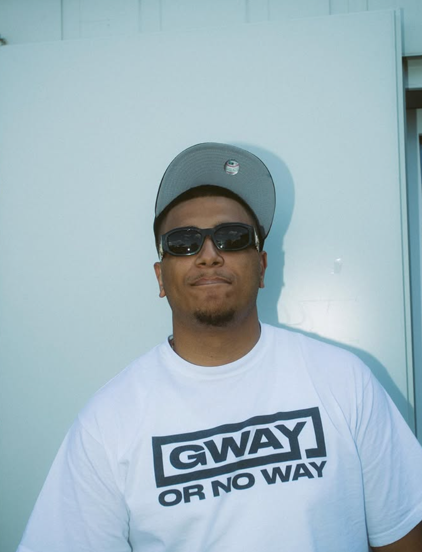 Man wearing sunglasses and a baseball cap stands against a light blue wall, wearing a white shirt with the text "GWAY OR NO WAY."