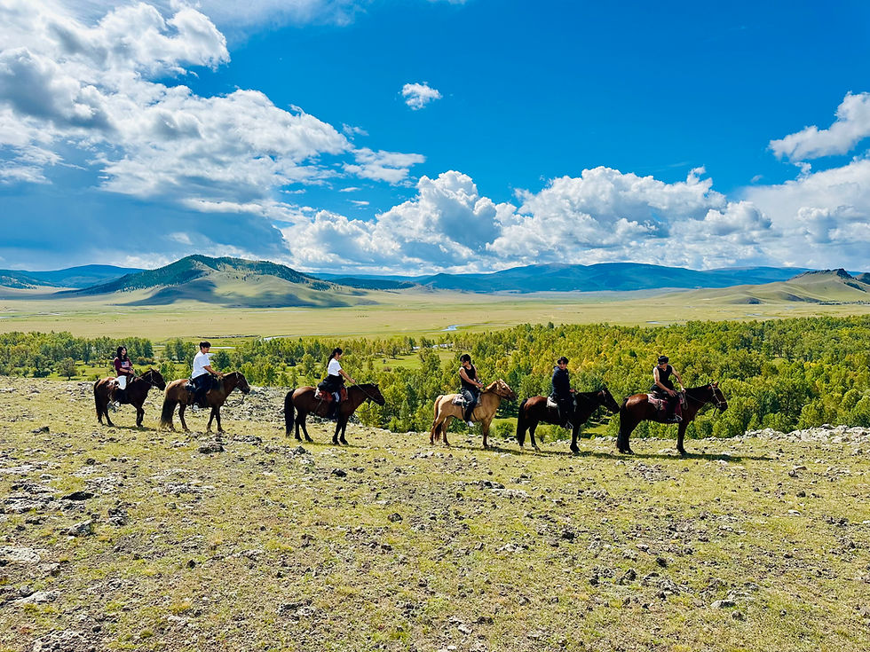 People riding horses across a grassy landscape under a blue sky with clouds. Green hills and distant mountains in the background.