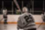 Hockey players in white jerseys on ice. Player with "29" prepares to swing stick. Other players in background. Dimly lit rink.