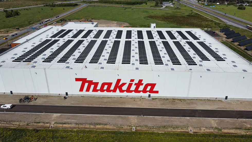 Aerial view of a large white building with "Makita" in red letters, featuring high-standard insulation and a roof covered with solar panels. The surrounding area includes grass, roads, and some vehicles.