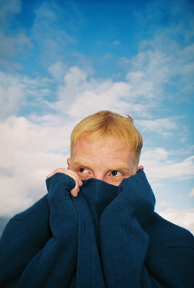 Wolf Henson with a blue sky background wearing a blue jumper pulled over his mouth