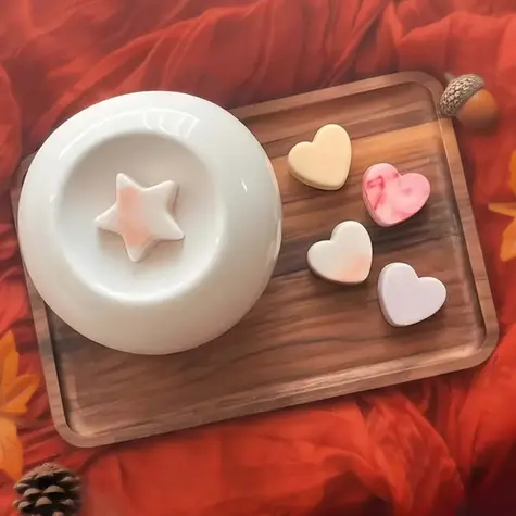 Autumn wax melts on a wooden board along side a ceramic white wax melt warmer. the board is sat upon an autumn orange soft fabric with pine cones and acorns.