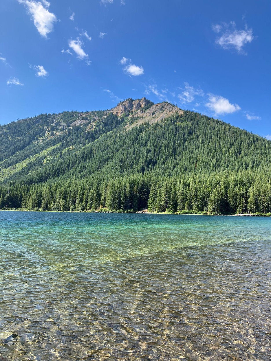 A forested mountain rises above a clear blue lake with a blue sky and whispy clouds above