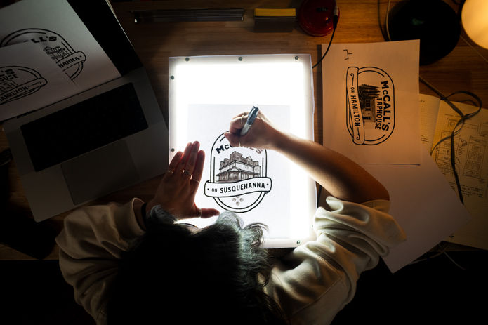 Artist hand-sketching logo of "McCall's Susquehanna" on a light box and papers.