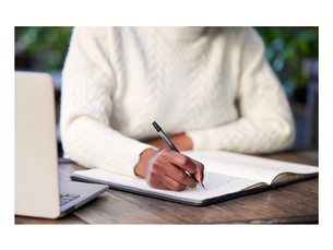 A person writes in a notebook next to an open computer