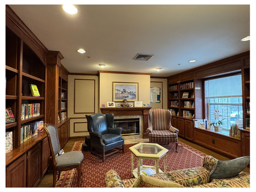 A picture of the mini-branch library at Waltonwood at University, a wood-paneled room with armchairs flanking a fireplace, and a cushioned couch and chair facing it. Books are arranged on built-in shelves on both sides of the room.