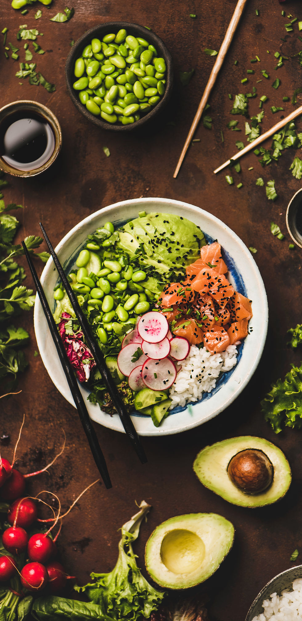 Salmon poke bowl with various vegetables, greens, sushi rice, soy sauce set among various fresh ingredients on a table
