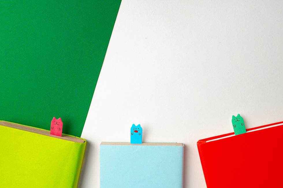 Three cut paper bookmarks with cat faces drawn on them stick out from the top of books.