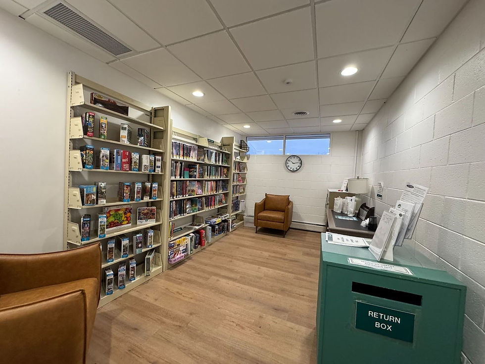 A picture of the mini-branch library at Avon on the Lake. A set of floor to ceiling shelves are filled with books and are flanked by two leather armchairs. A return box with library information on top is in the foreground of the photo.