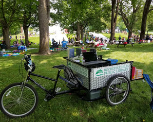 A bike with an attached trailer is parked in a busy park. The trailer has the RHPL logo on its side.
