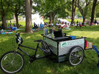 A bike with an attached trailer is parked in a busy park. The trailer has the RHPL logo on its side.