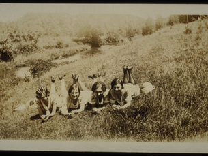 Sepia-toned historical photograph of four women lying on their stomachs on a hill in the grass with their feet up, smiling at the camera.