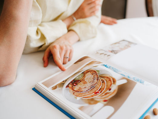 An arm and hand rest on a counter in front of a cookbook that is open to a page with a picture of pancakes.
