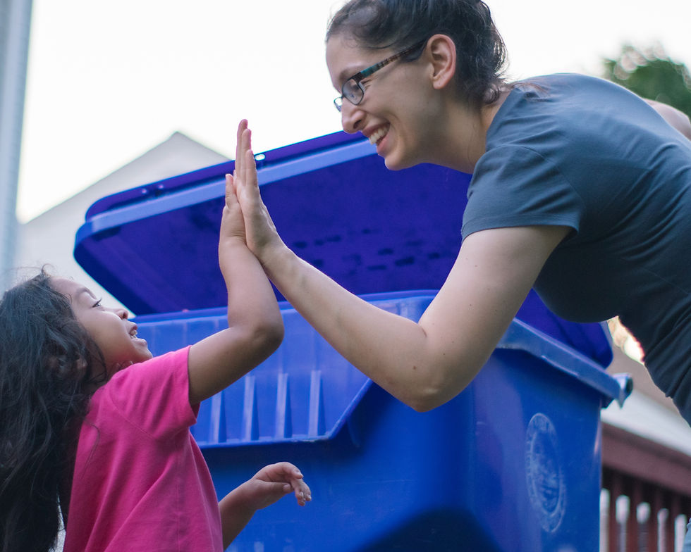 A monther and child high-five in front of a large, wheeled recycling bin.