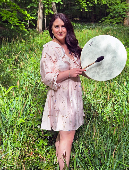 Alessandra steht im Wald mit einer Schamanischen Trommel und einem sommerlichen Kleid