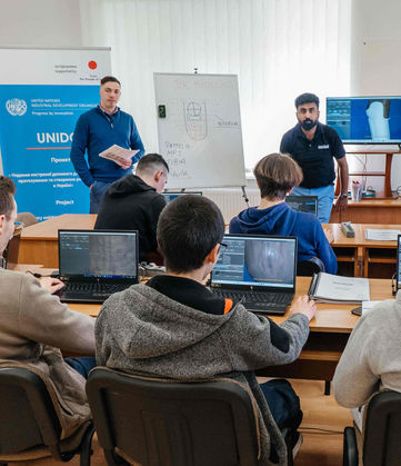 Group of students in a computer class with instructors, UN logo.