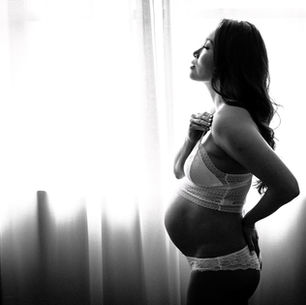Black and white maternity boudoir photograph of a pregnant woman in lace lingerie standing by a window, softly lit, celebrating pregnancy and feminine strength.