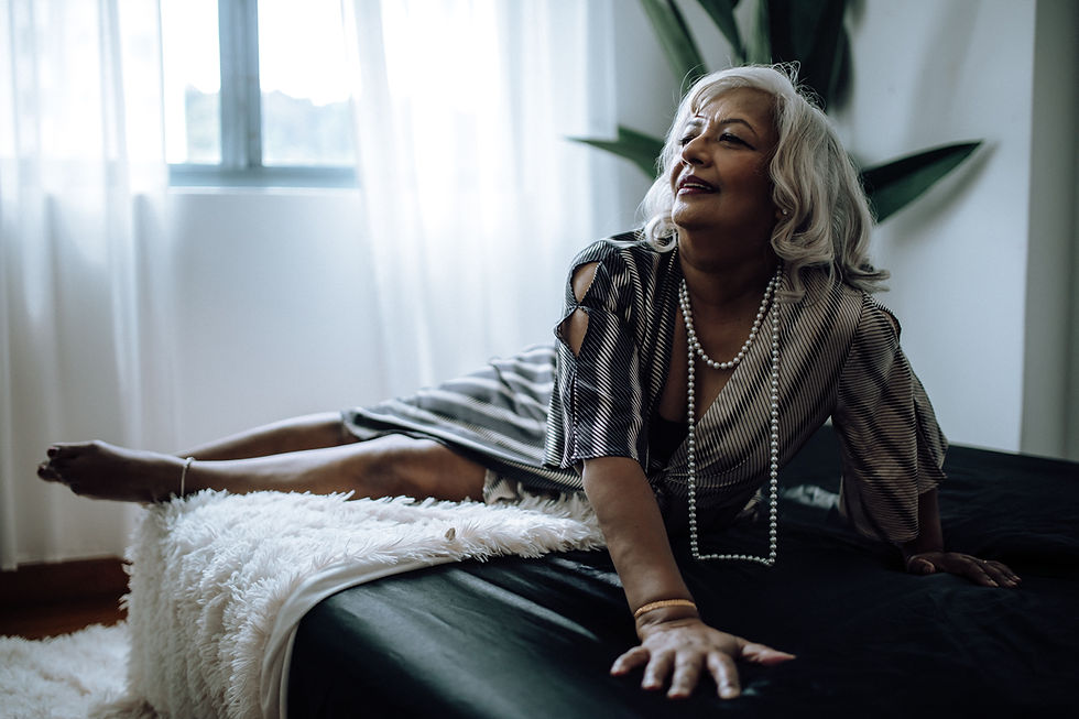 Elderly woman in a striped dress and pearls lounges on a bed, smiling. Bright room with plants and soft natural light through the window.