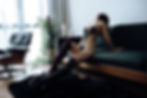 Woman in black lingerie and tall boots poses on a green sofa, back arched. Room with chair, plants, and soft lighting. Elegant mood.