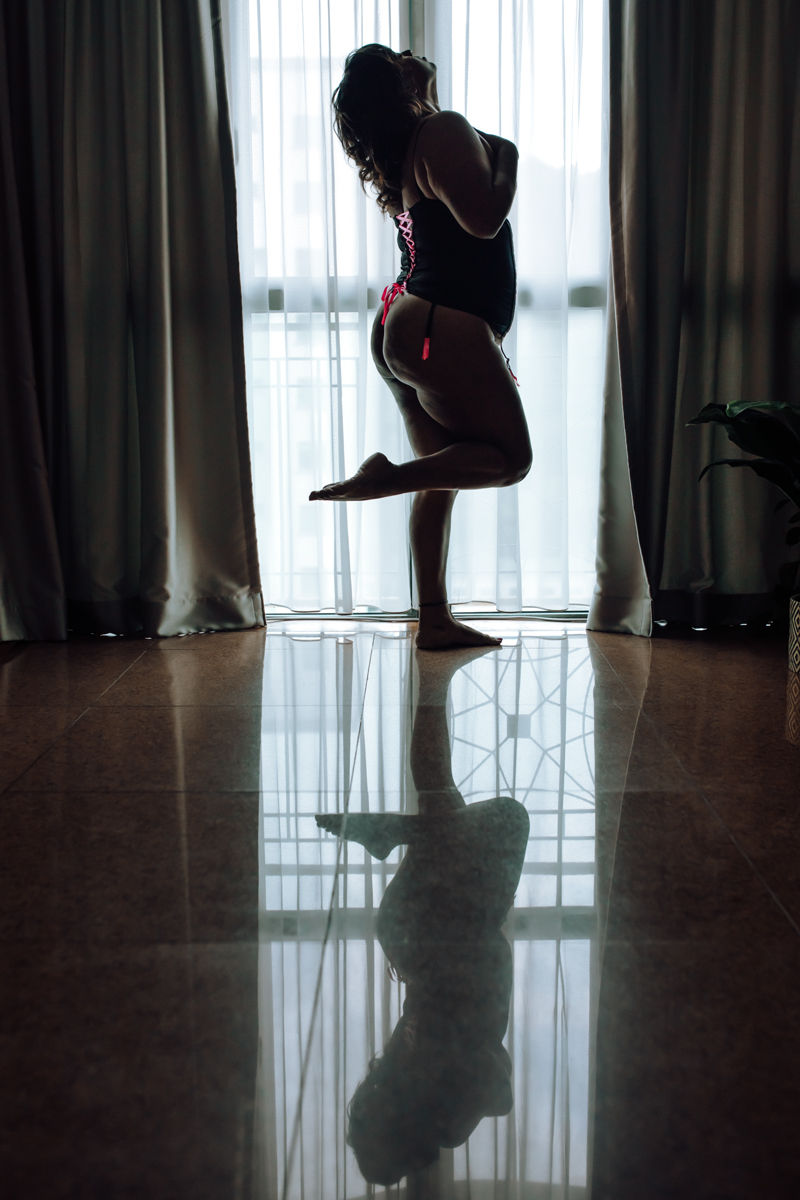 Silhouette of a woman in lingerie poses by a large window with sheer curtains; her reflection is visible on the polished floor.