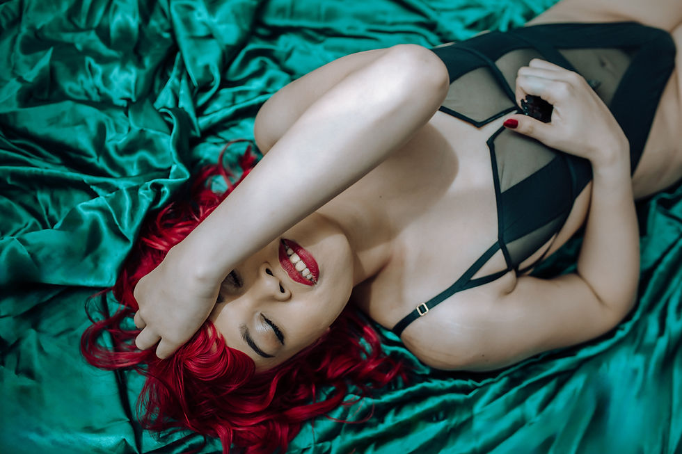 Red-haired person in black lingerie lies on emerald green satin, smiling with eyes closed, hand resting on forehead, relaxed mood.