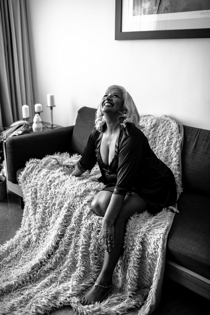Woman in a black robe sitting on a sofa with a fluffy blanket, smiling upwards. Candles and framed art in background. Black and white.