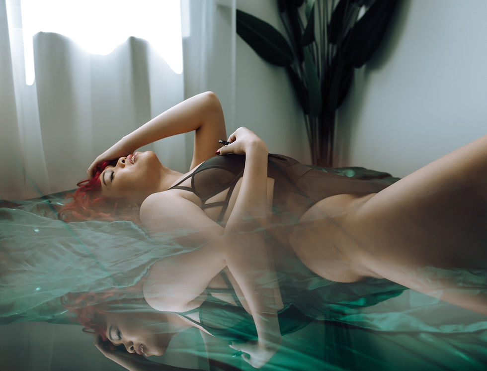 Person in black lingerie lying on a bed with teal sheets, hand in hair, relaxed expression. A plant and sheer curtains in the background.