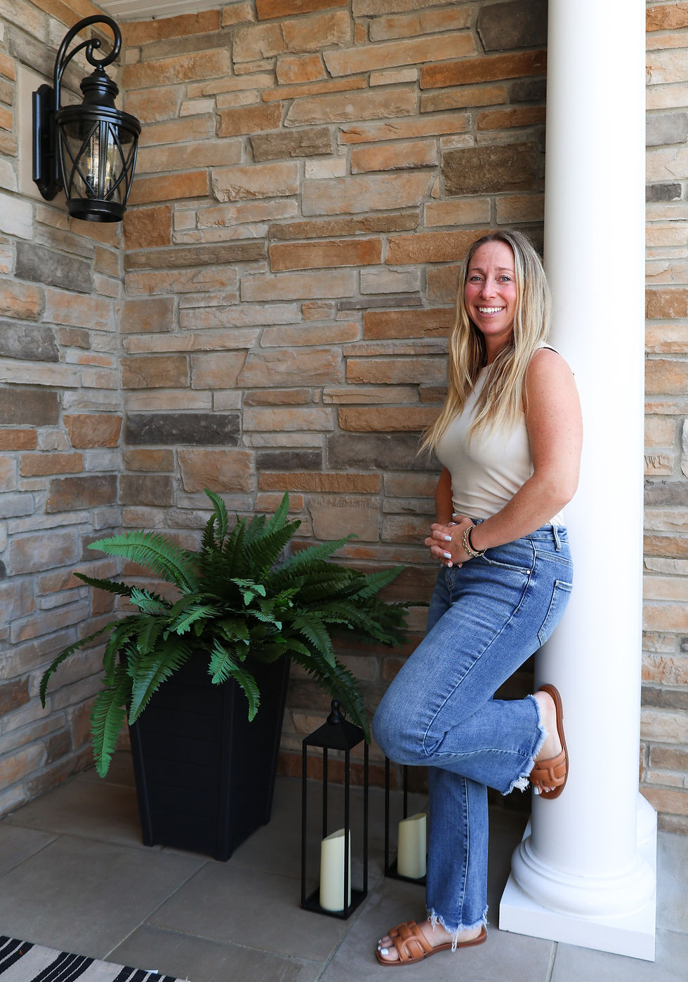 Jennifer DeVito the owner of Evolution leaning against her home in jeans and a tank top with plans and a stone wall.