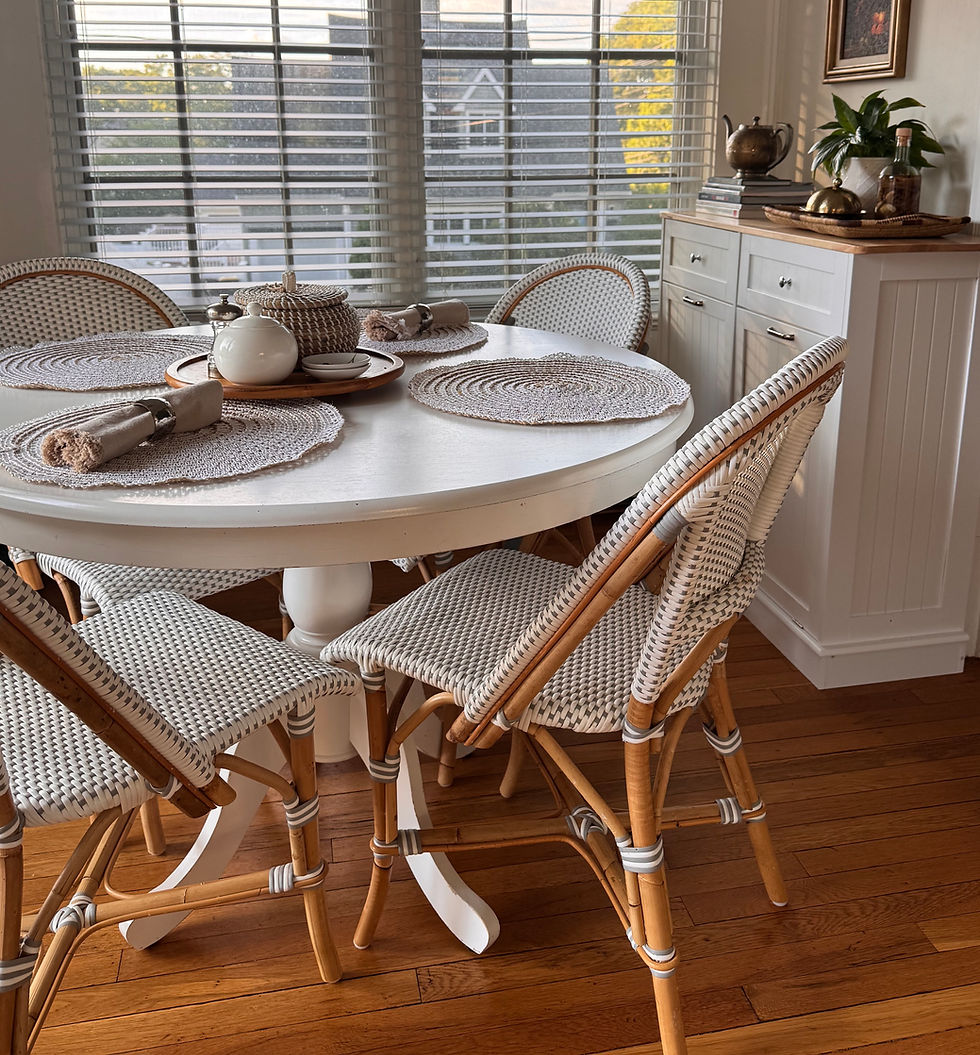 Sunlit dining nook with round white table, woven chairs, placemats, and tea set by window blinds; warm, calm, coastal feel.