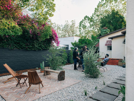 People socializing in a garden with wooden chairs and potted plants. Vibrant pink flowers and greenery cover a wall, creating a relaxed vibe.